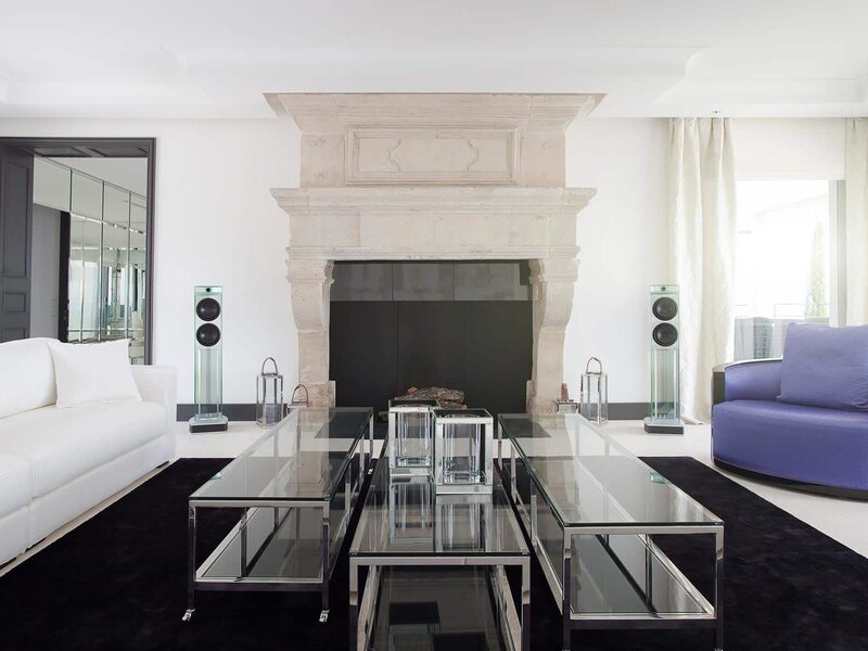 A white salon with slim glass-cabinet loudspeakers flanking a stone fireplace.